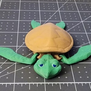 May include: A green and brown 3D printed turtle figurine with blue eyes. The turtle is lying on its belly with its legs outstretched.