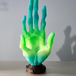 May include: A sculpted hand figurine with a gradient of green and teal hues. The hand is mounted on a dark brown, circular base. The fingers are elongated and slightly curved, creating a unique artistic piece.