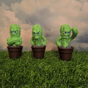 May include: Three green cactus figurines with angry expressions, each in a brown pot, sitting on a green grassy surface.