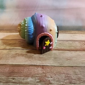 May include: A whimsical, colorful seashell house with a brown door and a yellow star. The shell is painted in pastel shades of blue, green, purple, and yellow. A pink arch frames the door.