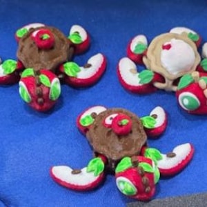 May include: Three turtle-shaped figurines crafted from clay, featuring shells made of brown textured circles or a beige pie-like structure. Each turtle has red apple-slice limbs with green leaves, and a red apple-shaped head with green eyes.