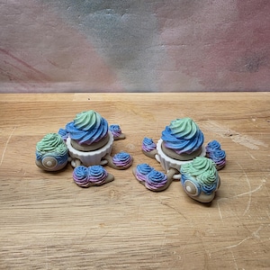 May include: Two turtle figurines with white shells and heads. Each turtle features a swirled, multi-colored frosting-like topping in shades of blue, purple, and green on its back. The heads and flippers are also decorated with the same colorful frosting.
