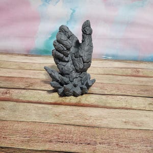 May include: A gray stone hand sculpture with a textured surface. The hand is raised with the thumb pointing up. The sculpture is on a wooden surface.