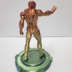 Puede incluir: Una figura de Iron Man impresa en 3D en un esquema de colores dorado y bronce. La figura est&aacute; de pie sobre una base circular verde con una superficie texturizada.