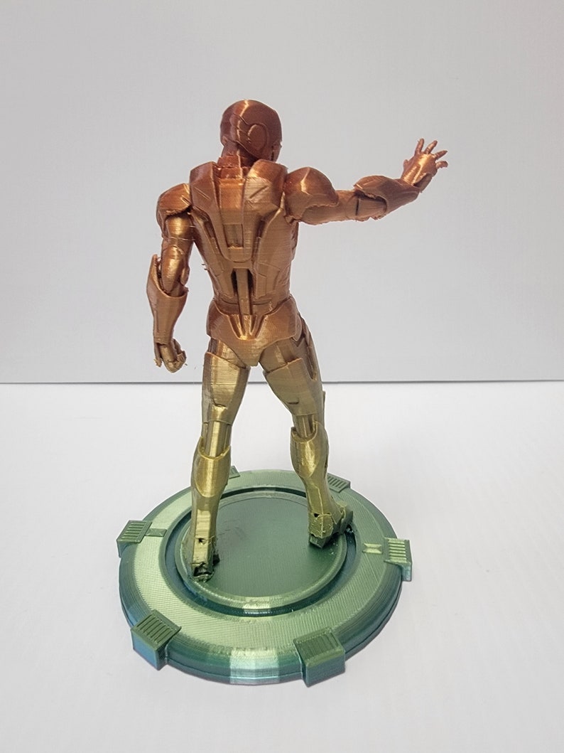 Puede incluir: Una figura de Iron Man impresa en 3D en un esquema de colores dorado y bronce. La figura est&aacute; de pie sobre una base circular verde con una superficie texturizada.