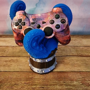 May include: A custom-designed video game controller with a galaxy-themed design, featuring shades of purple, pink, and orange. The controller is supported by a blue, textured base and a brown cylindrical stand with silver accents.