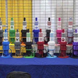 May include: A collection of colorful, oversized spark plug replicas with various brand logos displayed on a blue table. The plugs feature a range of colors, including red, blue, green, and black, and are arranged in rows against a yellow grid background.
