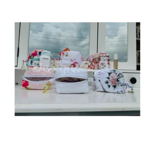 May include: Five small zippered pouches with floral patterns. The pouches are white with different floral designs. Two of the pouches are pink and white with floral patterns. One pouch is white with a floral pattern. One pouch is white with a floral pattern and a black and white design. The pouches are on a white surface.