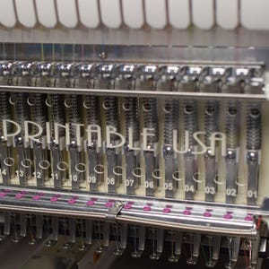 May include: Close-up of a silver embroidery machine with 15 needles, each with a small pink bobbin. The machine has a white label that reads "PRINTABLE USA".