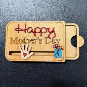 May include: Wooden Mother's Day card with the words "Happy Mother's Day" in red and brown. The card features a hand with a heart and a vase of flowers. The card has a sliding mechanism.