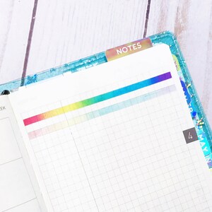 May include: An open planner with a rainbow color gradient across the top of the page. The planner has a gridded page on the right and a blank page on the left. The left page has the text "15th WEEK" at the top. The right page has a tab that says "NOTES" at the top.