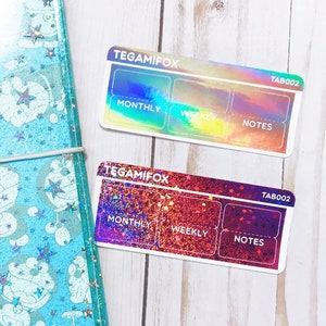 Peut inclure: Un agenda transparent bleu clair avec un motif d'étoiles et de nuages et un charme argenté. Deux onglets d'agenda irisés avec le texte "TEGAMIFOX" et "TAB002" sont visibles. Les onglets sont étiquetés "MENSUEL", "SEMAINE" et "NOTES".