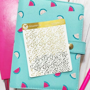 Puede incluir: Una agenda azul turquesa con estampado de sandía y una pegatina dorada con confeti negro y dorado. La agenda tiene una cremallera dorada y un bolígrafo rosa está a su lado.