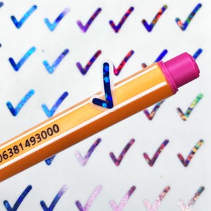 Puede incluir: Una hoja de pegatinas de marca de verificación holográficas con un acabado iridiscente azul, rosa y morado. Un lápiz amarillo con una goma de borrar rosa se coloca encima de las pegatinas. El lápiz tiene el número 4006381493000 impreso en él.