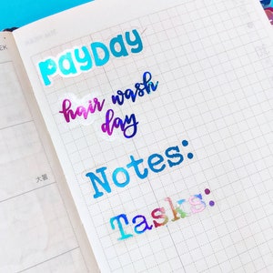May include: A close-up of a planner with colorful stickers that say "Payday", "Hair Wash Day", "Notes:", and "Tasks:".