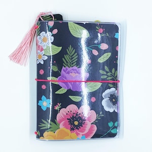 May include: A floral-patterned journal with a dark background, featuring colorful flowers in pink, purple, yellow, and blue. A pink elastic band secures the journal, and a matching tassel hangs from the top. The journal is encased in a clear plastic cover.