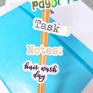 Può includere: Una matita con quattro adesivi colorati attaccati. Gli adesivi dicono "payDo", "task", "Notes" e "hair wash day".