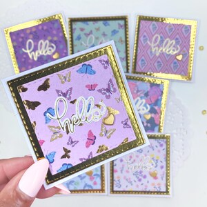 Op de afbeelding: Een verzameling vierkante wenskaarten met vlinder- en bloempatronen. Elke kaart heeft een gouden rand en het woord "hello" in sierlijke letters. De kaarten zijn in tinten paars, roze en blauw, met gouden accenten.