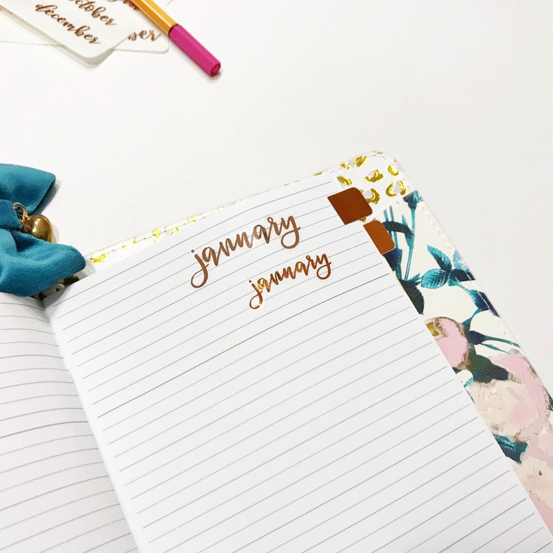 May include: An open journal with lined pages, a floral patterned cover, and gold accents. The word "january" is written in gold lettering on the page.