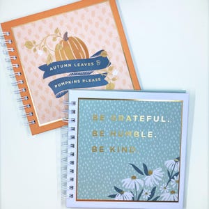 May include: Two spiral-bound notebooks. One has an orange cover with a pumpkin graphic and the text "AUTUMN LEAVES & PUMPKINS PLEASE." The other has a blue cover with the text "BE GRATEFUL. BE HUMBLE. BE KIND." and white floral accents.