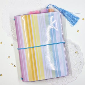 May include: A rectangular notebook with a glossy cover featuring vertical stripes in shades of yellow, orange, pink, and blue. The notebook is secured with a bright blue elastic band and includes a matching light blue tassel attached to the top.