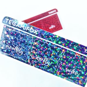 May include: A sheet of planner stickers with a holographic blue background covered in colorful stars. The stickers have labels that say "monthly", "weekly", and "notes". The brand name "TEGAMIFOX" is printed at the top.