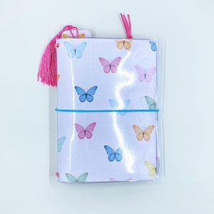 May include: A clear, plastic-covered journal with a white background and colorful butterfly pattern. The journal is secured with a blue elastic band and features a pink tassel and a small tag. The butterflies are in shades of blue, pink, orange, and green.