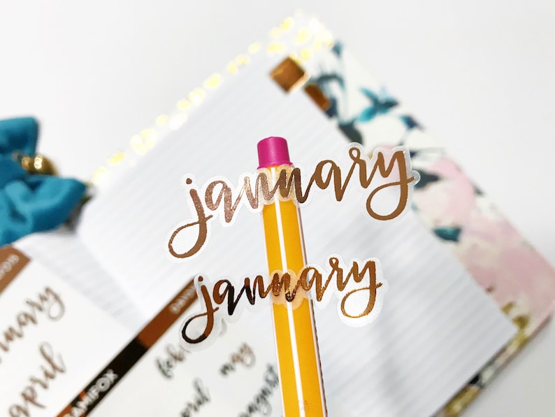 May include: Two gold foil stickers with the word "january" written in cursive script. The stickers are on a yellow pencil with a pink eraser.