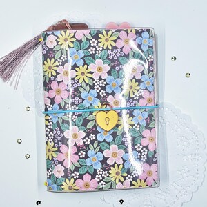 May include: A rectangular journal with a glossy cover featuring a dark background and a dense pattern of pink, yellow, and blue flowers with green leaves. A light blue elastic band wraps around the center, holding a yellow heart-shaped padlock.