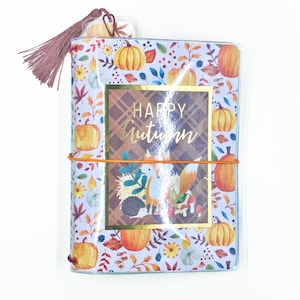 May include: A decorative journal with a fall theme. The cover features pumpkins, leaves, and a fox illustration. The words "HAPPY Autumn" are printed on the front. An orange elastic band holds the journal closed, and a tassel is attached to the top.
