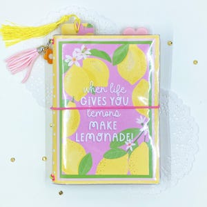 May include: A notebook with a pink cover featuring yellow lemons and the text "when life gives you lemons make lemonade!". It has a yellow and pink tassel, a pink elastic band, and a flower charm.
