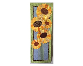 Könnte beinhalten: Ein Wandbehang mit blauem Hintergrund und fünf gelben und braunen Sonnenblumen. Die Sonnenblumen sind vertikal angeordnet und von einem grünen Rand umgeben.