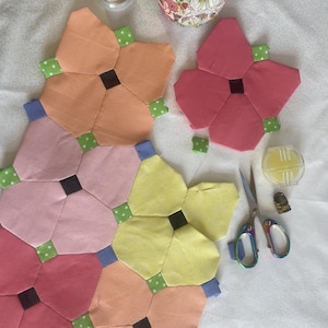 Puede incluir: Un primer plano de una colcha de retazos hecha de cuadrados de tela en varios tonos de rosa, naranja y amarillo. La colcha está hecha de bloques con forma de flor con acentos verdes. La colcha se está trabajando con herramientas y suministros de costura, incluyendo un alfiletero, tijeras e hilo.