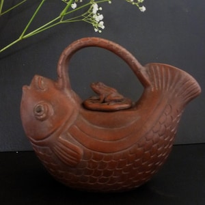 Puede incluir: Una tetera de cerámica marrón con forma de pez con una rana en la tapa. La tetera tiene un diseño detallado de escamas y aletas.