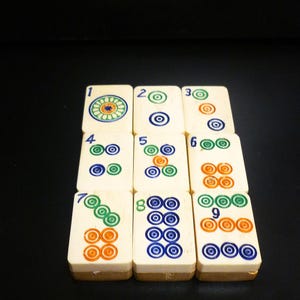 Op de afbeelding: Een set ivoorkleurige Mahjong-tegels, elk met unieke symbolen en cijfers in groen, blauw en oranje. De tegels zijn gerangschikt op een donkere ondergrond, wat het ingewikkelde ontwerp en het culturele erfgoed van het spel laat zien.