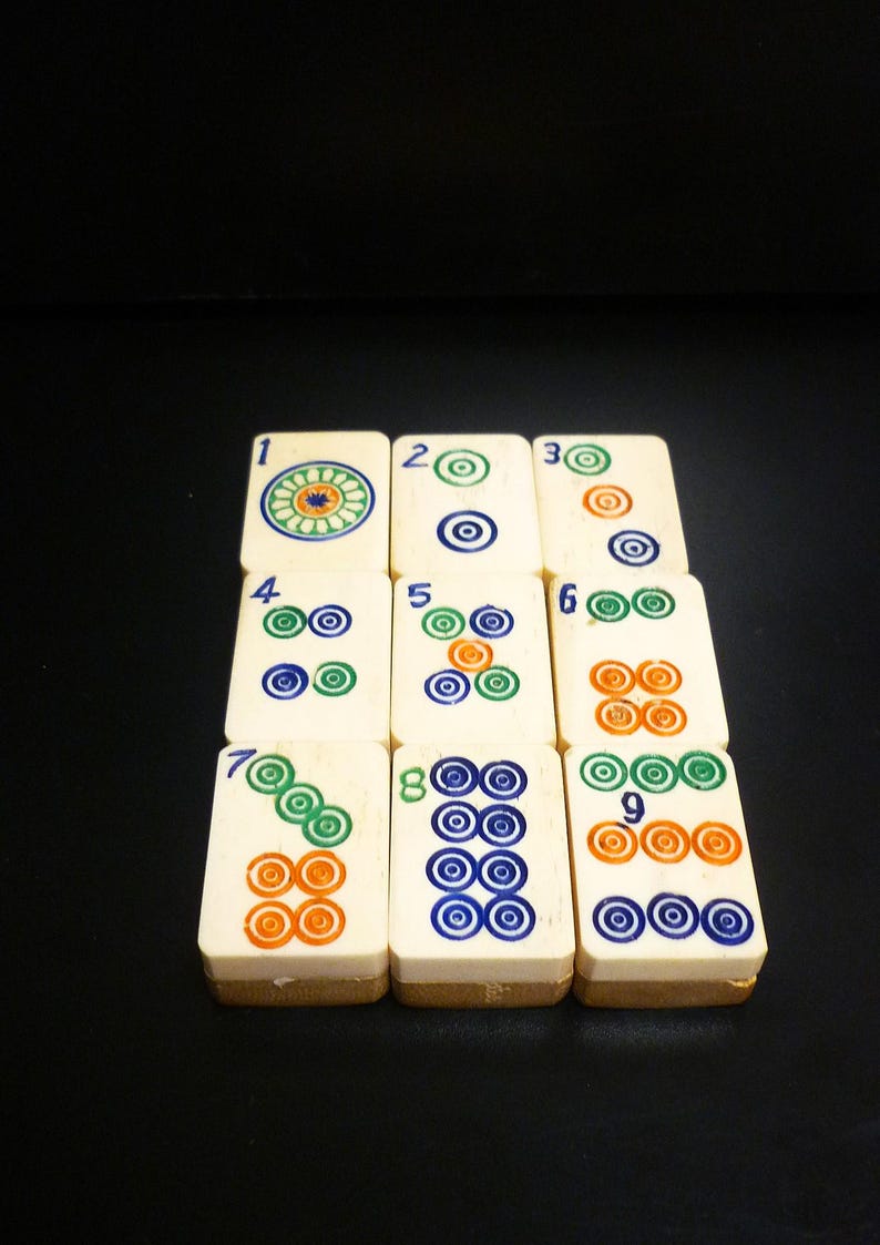 Op de afbeelding: Een set ivoorkleurige Mahjong-tegels, elk met unieke symbolen en cijfers in groen, blauw en oranje. De tegels zijn gerangschikt op een donkere ondergrond, wat het ingewikkelde ontwerp en het culturele erfgoed van het spel laat zien.