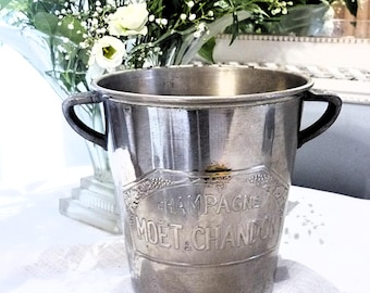 May include: A silver champagne bucket with two black handles. The bucket has the words "CHAMPAGNE MOET & CHANDON" embossed on the front. The bucket is sitting on a white surface, with a vase of white flowers in the background.