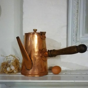 Peut inclure: Une cafetière en cuivre avec une poignée en bois et un bec verseur. La cafetière a une finition martelée et un couvercle à charnière.