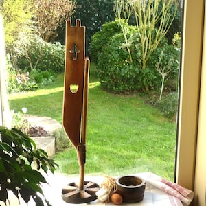 May include: A tall, wooden decorative piece with a cross and heart-shaped cutouts. The piece stands on a round base. A mortar and pestle, garlic, and an egg are on a white surface in front of a window overlooking a green yard.
