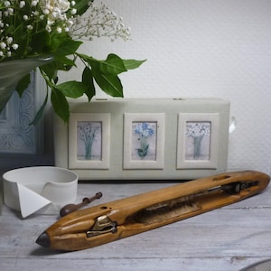 Könnte beinhalten: Ein hölzerner Spulen-Shuttle mit weißem Garn, eine weiße Box mit drei gerahmten Blumenprints und eine weiße Keramikschale auf einer Holzoberfläche.