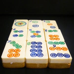 Op de afbeelding: Een set ivoorkleurige Mahjong-tegels, elk met unieke combinaties van gekleurde cirkels en cijfers. De tegels zijn in een netjes patroon gerangschikt tegen een donkere achtergrond, wat het ingewikkelde ontwerp van het spel laat zien.