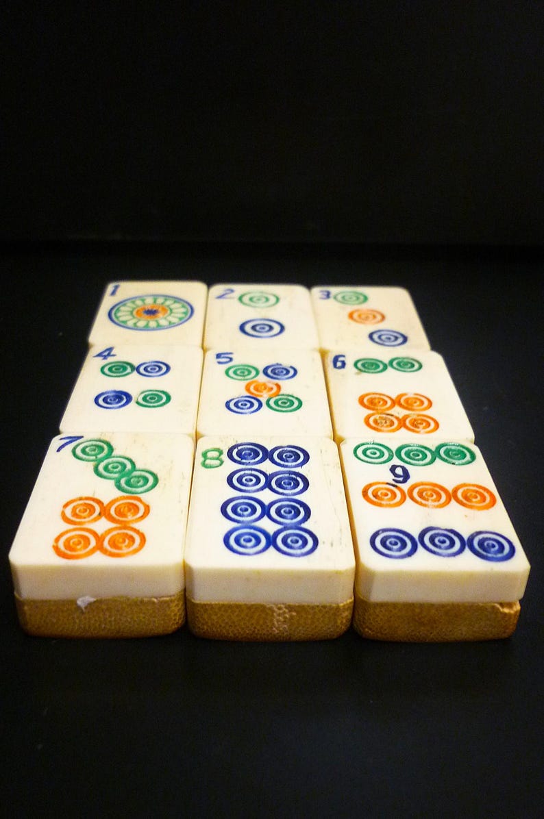 Op de afbeelding: Een set ivoorkleurige Mahjong-tegels, elk met unieke combinaties van gekleurde cirkels en cijfers. De tegels zijn in een netjes patroon gerangschikt tegen een donkere achtergrond, wat het ingewikkelde ontwerp van het spel laat zien.