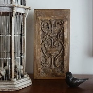 May include: A rectangular wooden panel featuring a carved relief of a central urn with symmetrical scrolling foliage and floral motifs. Beside it stands a tall, weathered wooden birdcage with wire bars and a small, dark, smooth bird figurine.