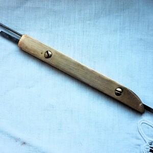 Op de afbeelding: Een vintage priem met een houten handvat en metalen lemmet. De priem heeft een lang, slank ontwerp met een punt en een plat, rechthoekig lemmet. Twee schroeven bevestigen het handvat. De priem wordt waarschijnlijk gebruikt voor het doorboren of markeren van materialen.