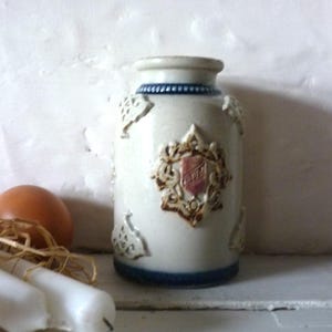 May include: A white ceramic jar with blue and brown accents. The jar has a decorative design with a crest and floral motifs. The jar is approximately 6 inches tall.