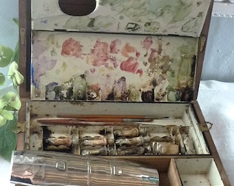 May include: A wooden artist paint box with a hinged lid featuring a built-in palette with dried paint residue. The interior contains several thin paintbrushes and multiple metal paint tubes. A large, polished silver-toned metal object rests inside.