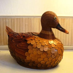 May include: A decorative duck figurine crafted from woven wicker and wooden scales. The duck features a brown head and neck, a dark beak, and a body covered in overlapping wooden scales in shades of brown and orange. A gold-colored eye is visible.