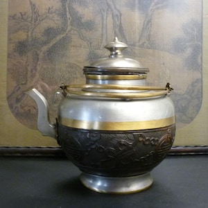 May include: A silver teapot with a gold band and intricate carvings. The teapot has a lid with a knob and a spout. The teapot is sitting on a wooden surface.