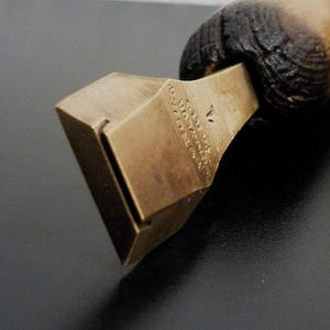 Op de afbeelding: Close-up van een vintage messing gereedschap met een houten handvat. Het messing deel heeft een geometrische vorm met een rechthoekige opening en gegraveerde tekst. De houten handgreep is donkerbruin en lichtbruin.