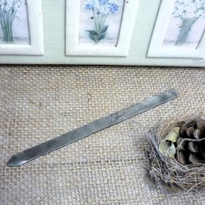 Op de afbeelding: Een lang, dun metalen gereedschap met een puntig uiteinde, waarschijnlijk voor knutselen of detailwerk, ligt op een getextureerd jute oppervlak. Een klein decoratief nest met een dennenappel en andere natuurlijke elementen bevindt zich op de achtergrond, samen met ingelijste bloemenkunst.